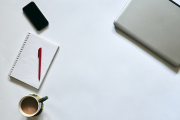 Flat-lay picture of office accessories, isolated on white background. Grey metallic laptop, white smart phone, red pen, cell notepad and gray cup of coffee drink on table. Work online. Stay home.