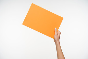 Cropped view of young woman holding in raised hand inclined piece of yellow paper while standing against white background. Human hands and signs concept
