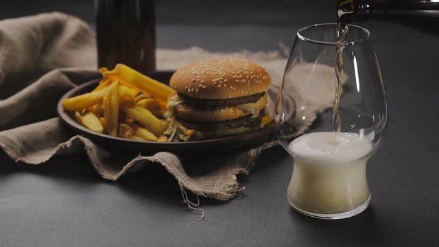 Pouring fresh and cold beer from dark bottle into glass with white foam on top. French fries on dark plate next to hamburger. Black table. Close up