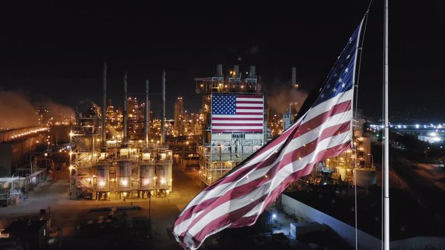 Aerial view over operating petroleum refinery at night. Brightly illuminated constructions of the huge modern plant. American flag waving on the wind is on the foreground. United States. 4K