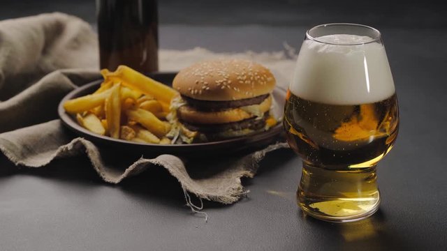 Pouring fresh craft beer from dark bottle into glass with white froth on top. French fries on dark plate next to hamburger. Black table. Close up