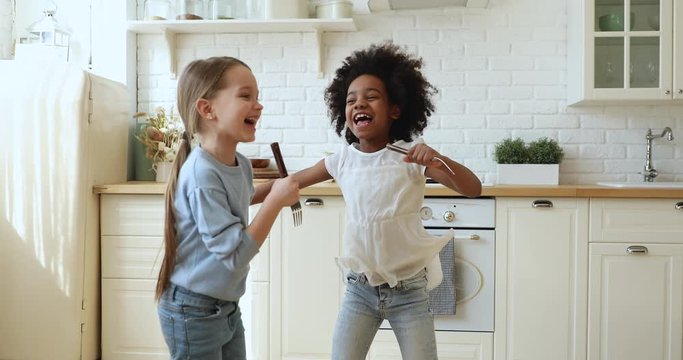 Happy cute african and caucasian small girls stepsisters or friends having fun playing together in kitchen. Two funny mixed race children dancing, jumping, singing in kitchenware microphones at home.