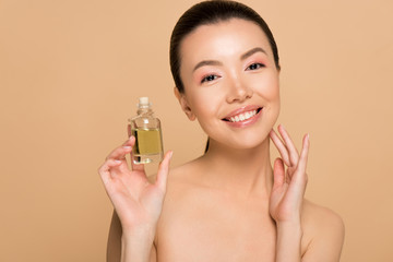 attractive cheerful nude asian girl applying oil on face isolated on beige