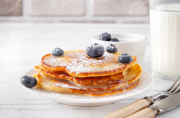 fried pancakes with  blueberries on a plate,   fork,  knife,  glass of milk on a white wooden