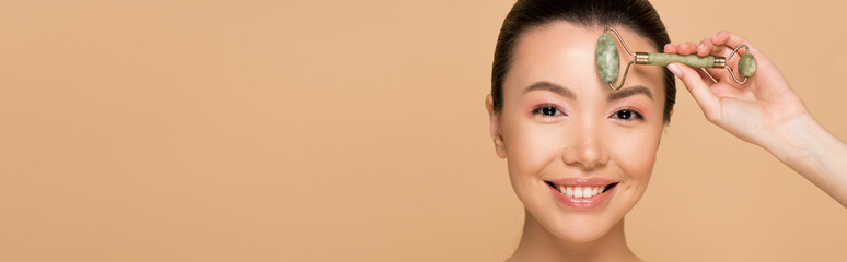 panoramic shot of beautiful smiling asian girl making massage of face with jade roller isolated on beige