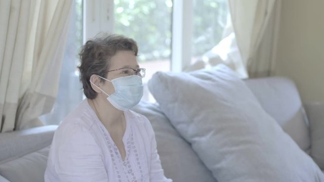 Happy beautiful older middle aged woman wearing protective mask relaxing sitting on sofa at home, stay at home health care COVID-19 pandemic situation.
