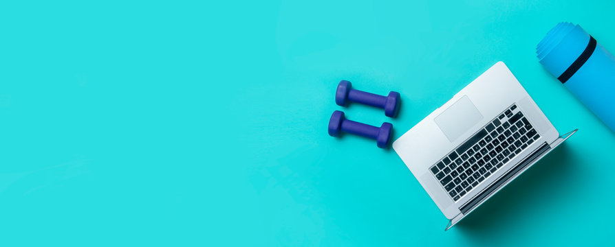 Blue dumbbells, gym mat and grey laptop on blue background. Online workout concept