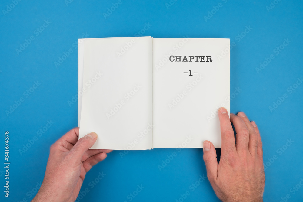 Fototapete top view of person fiipping open a book with text chapter 1 on first page against blue background