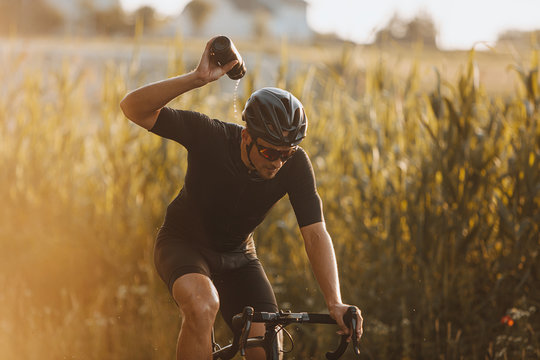 Professional athlete wearing sport clothing, black helmet and protective glasses splashing fresh water from bottle while sitting on bicycle. Concept of refreshing and exercise routine