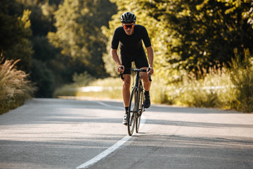 Strong sportsman with athletic body wearing black sport clothing, helmet and sunglasses practicing in cycling on fresh air. Concept of healthy hobby and regular trainings - obrazy, fototapety, plakaty