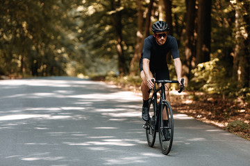 Naklejka premium Professional cyclist in sport clothing and black helmet riding bike on paved road among summer forest. Bearded athlete in protective eyeglasses training outdoors.