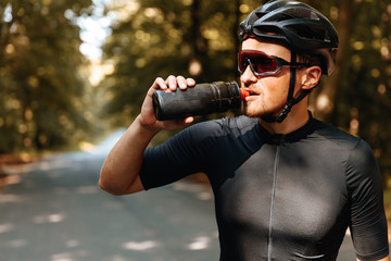 Portrait of exhausted sportsman in black helmet and mirrored glasses drinking cold water from sport...