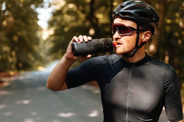 Fotobehang Portrait of exhausted sportsman in black helmet and mirrored glasses drinking cold water from sport bottle while standing in green forest. Mature man having break during long ride. © Tymoshchuk