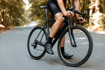 Close up of mature man in sport clothing standing on paved road among forest with black bicycle. Professional sportsman having rest between hard training outdoors.