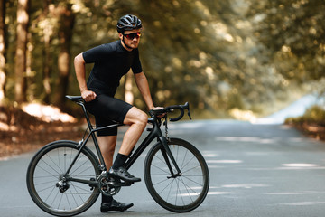 Sporty bearded man in cycling clothes, protective helmet and mirrored glasses posing on camera with black bike outdoors. Concept of sport and active lifestyle