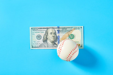 Baseball ball and banknote of 100 dollars on blue background. Purchasing sport accessories. Concept of corruption in sport
