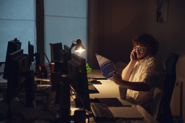 Talking on phone. Man working in office alone during COVID-19 quarantine, staying to late night. Young businessman, manager doing tasks with smartphone, laptop, tablet in empty workspace.