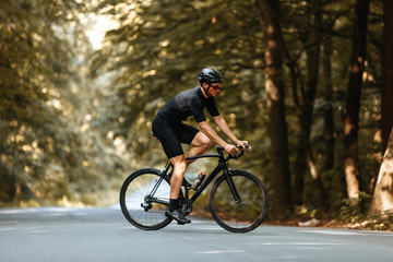 Fototapeta na wymiar Side view of active man in sport clothing riding black bike with beautiful green trees around. Bearded cyclist in protective helmet and eyewear training regularly to prepare for races.