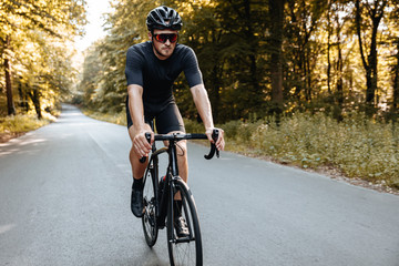 Professional cyclist in sport clothing and protective helmet actively riding bike on paved road among green trees. Strong man in mirrored glasses workout on nature.