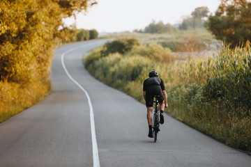 Back view of strong male cyclist with athletic body shape riding bike at the paved road among trees...