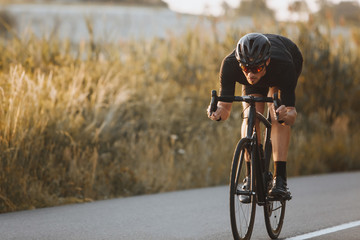 Active professional sportsman cyclist wearing black sports outfit, helmet and glasses riding bike at the paved road outdoors. Strong man improving skills and getting ready for cycling competition - obrazy, fototapety, plakaty