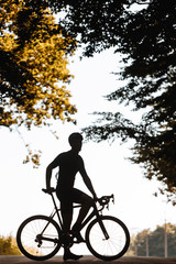Strong man in silhouette wearing cycling clothes and helmet, posing on camera with bike among green...