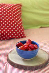 Bowl of fresh strawberries on a bed with striped bedding. Selective focus.