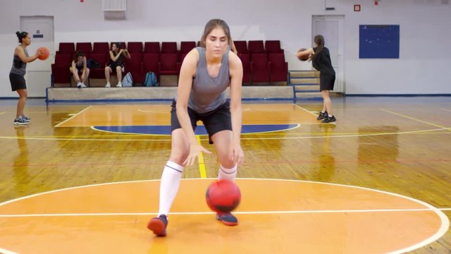 Young professional female athlete in sportswear looking at camera and dribbling a basketball on indoor court while her teammates training in the background