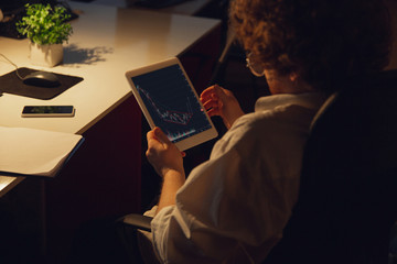 Analyzing graphs. Man working in office alone during COVID-19 quarantine, staying to late night. Young businessman, manager doing tasks with smartphone, laptop, tablet in empty workspace.