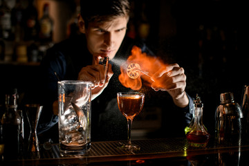 Young bartender holds tweezers with slice of citrus over glass sprinkles and sets it on fire.