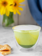 Hot matcha latte in a glass cup on a light gray table, next to crackers and a vase with blooming sunflowers. Close-up, shallow depth of field.