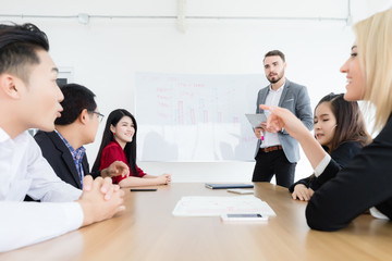 The male supervisor is explaining the company's performance charts to his employees and other employees in the office meeting room.