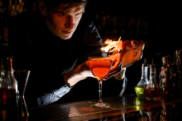 Young male bartender sprinkle to alcoholic cocktail with piece of citrus zest and make fire