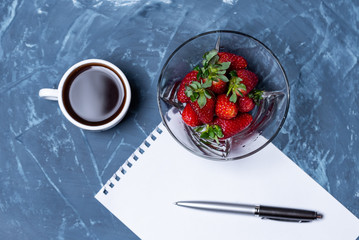 Strawberries in a transparent plate, a cup of coffee and a notebook page