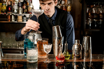Young bartender holds clean glass in his hand and looks at it.