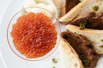 Red caviar in a transparent glass on a background of bread and a white plate. Above view.