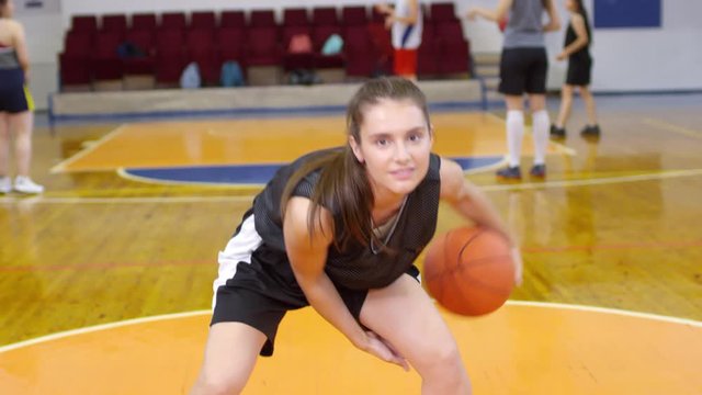 Young pretty girl in sports uniform looking at camera and performing tricks with basketball while training with female team on indoor court