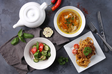 Complex dinner. Fresh vegetable salad, chicken broth soup, bolognese pasta on white plates and a pot of tea on a dark grey background. Fresh herbs, garlic and chili pepper, fork and knife. Top view