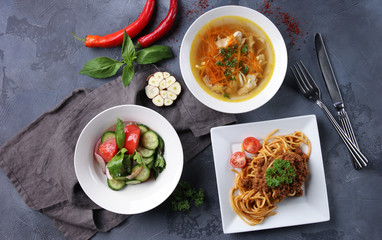 Complex dinner. Fresh vegetable salad, chicken broth and bolognese pasta on white plates on a dark grey background. Fresh herbs, garlic and chili pepper, fork and knife. Top view. Background image