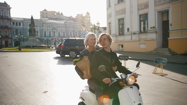 Happy young hipster couple riding a vintage scooter in old city center with grocery paper bag and talking to each other, tracking shot, slow motion