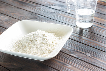Flour in white plate, measuring glass with water. Sun's rays on table.