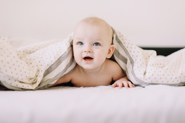 Adorable baby learning to crawl in white  bedroom. Cute funny baby lying under the blanket on bed. Little child doing tummy time