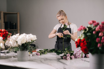 Pretty young woman florist wearing apron cutting fresh rose to bouquet at the table on white background. Concept of working with flowers, floral business.