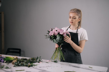 Professional young woman florist wearing apron making floral composition from fresh roses at the table on white background. Concept of working with flowers, floral business.