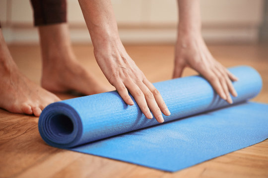Mature Woman Folding Blue Yoga Or Fitness Mat Before Or After Working Out At Home In Living Room. Healthy Life In Covid-19 Time Lockdown. Focus On Hand.
