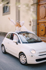 Beautiful bride standing up and waving in a convertible in front of the Medici Villa of Lilliano Wine Estate, Tuscany, Italy.