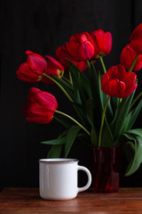 White coffee cup with amazing blooming red tulips bouquet on black background. Dark moody low key minimalism style flowers and hot drink mug photo banner. Stylish greeting card copy space.Good morning