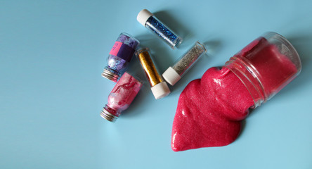 Pink slime flows out of a container next to small jars of colored paints and glitters on a blue background