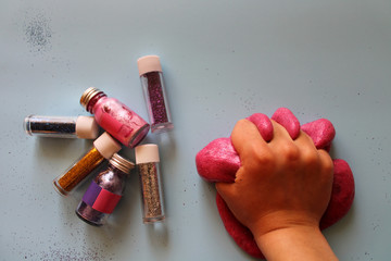 Child’s hands play with pink slime on a blue background next to multicolored paints and glitters