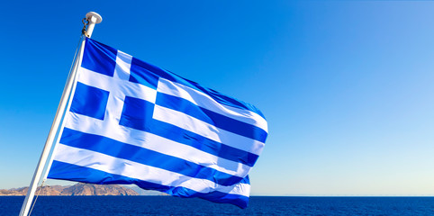 waving Greek flag against blue sky and the coastline of the island
